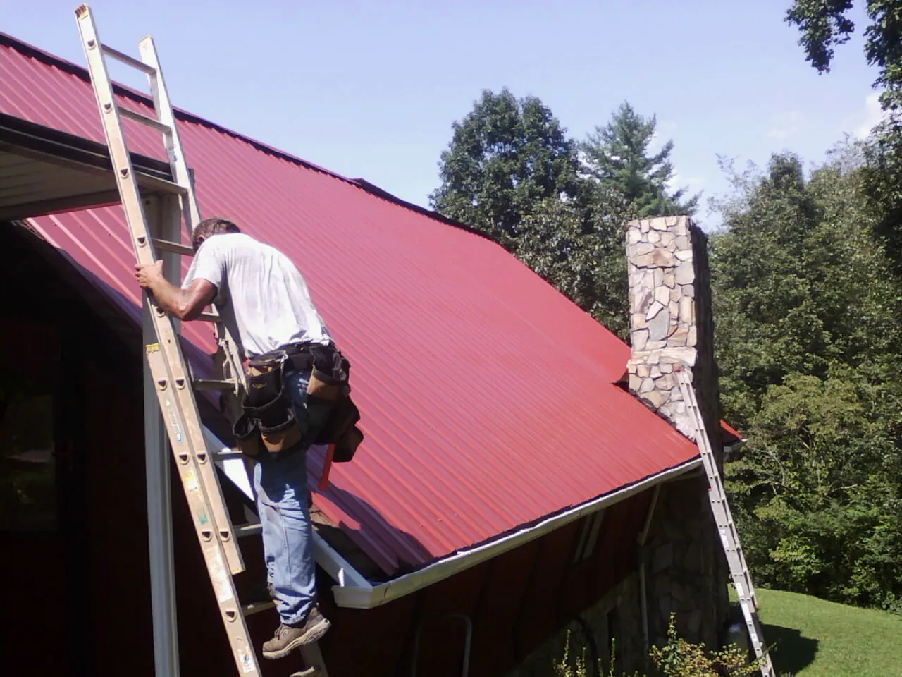 Roofer on a ladder beside a red metal roof and stone chimney — Soffit & Fascia Repair by Roofing Brothers in Wallops Island